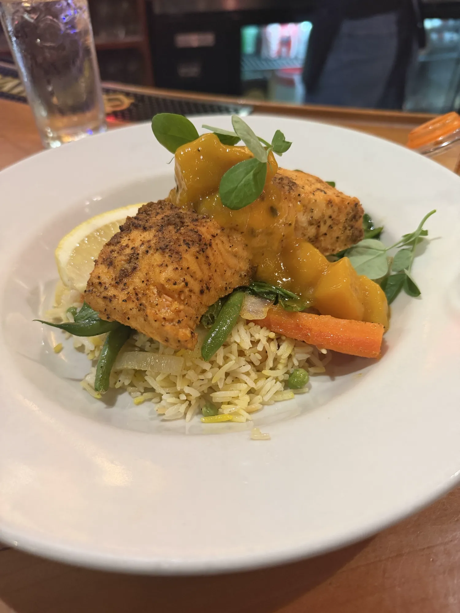 Broiled salmon at Ama Kitchen — two fillets dusted with black pepper, draped in saffron-mango chutney, set over jasmine rice, snap peas, and carrot, with a lemon wedge and pea-shoot garnish.