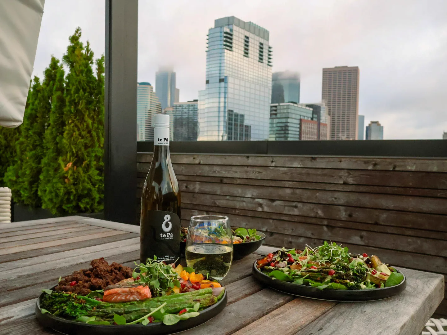Two plates on a weathered rooftop table beside a bottle of te Pā Sauvignon Blanc and a half-poured stem glass, downtown Minneapolis skyline behind in the late light.