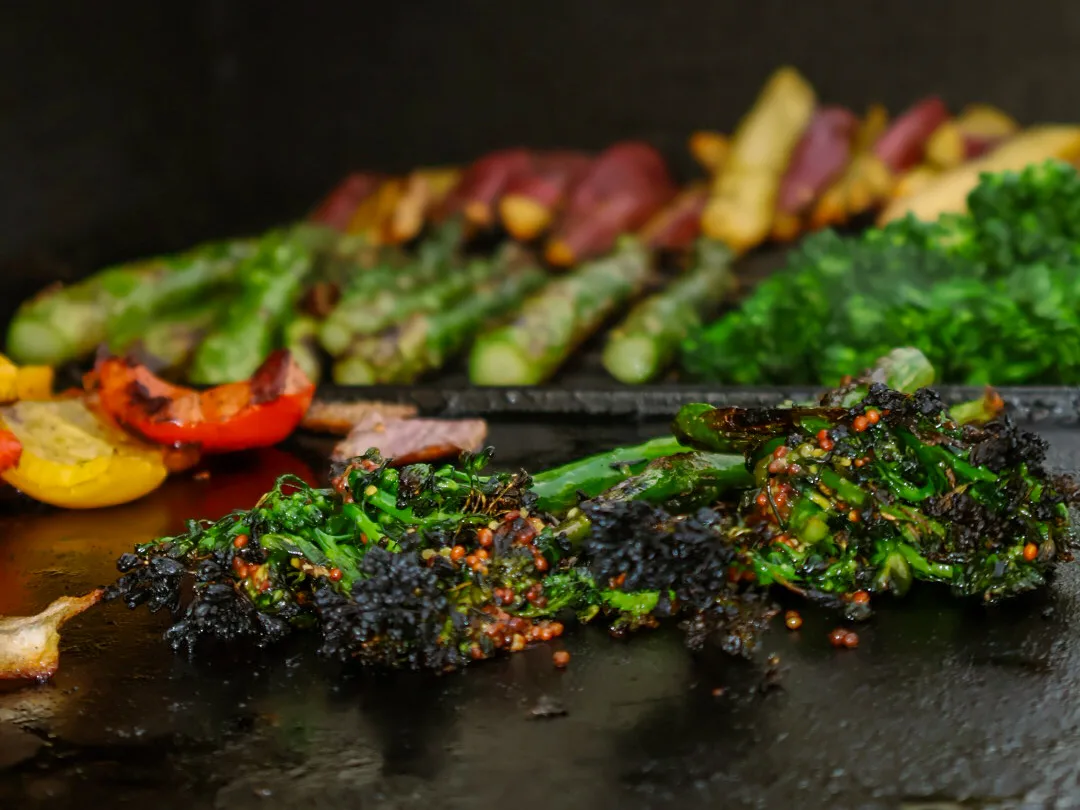 Charred broccolini coming off the hot grill onto a black plancha, blackened florets and bright green stems, the moment before the warm mustard toss.