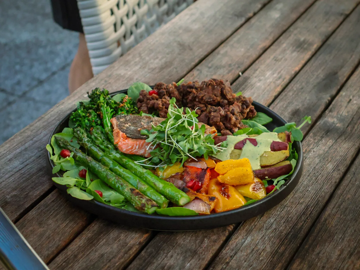 Single dark plate on weathered wood: wild salmon centered, roasted sweet potato, charred broccolini, asparagus, peppers, watercress, micro-greens, tahini-cilantro drizzle, pomegranate seeds.