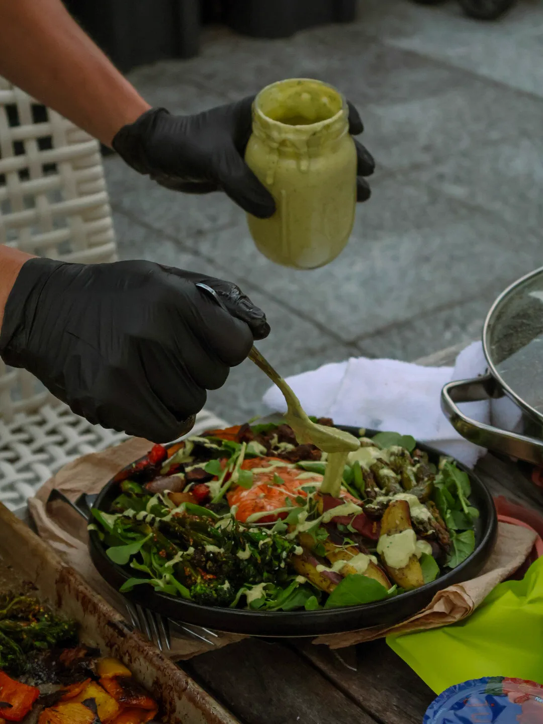 Black-gloved hands pour tahini-cilantro-lemon sauce from a green-streaked mason jar onto a finished salmon plate on the rooftop, the ribbon mid-pour catching the late light.
