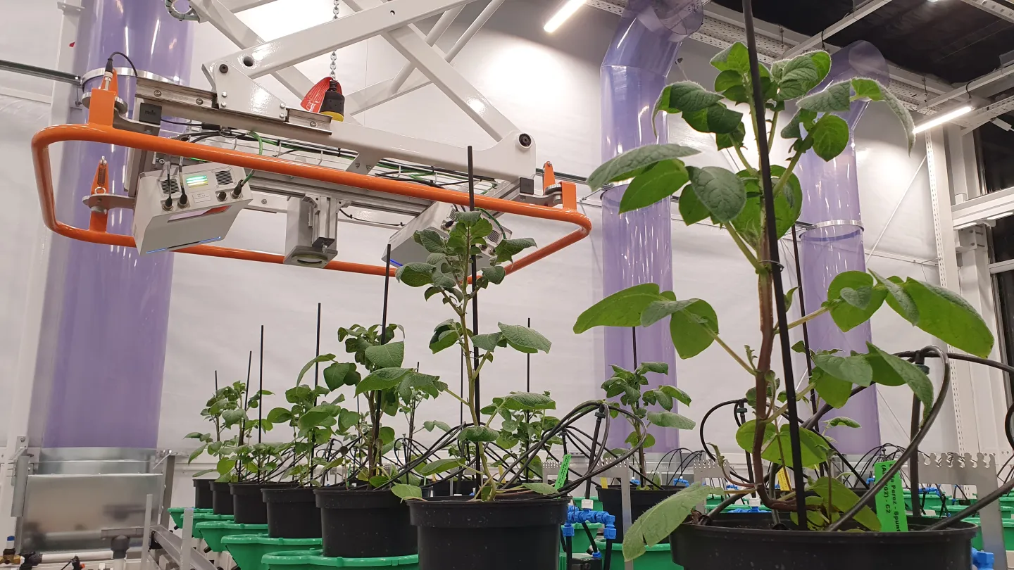 NPEC at WUR — an overhead orange gantry-mounted multispectral sensor array scanning rows of potted plants inside a sealed indoor growth room.