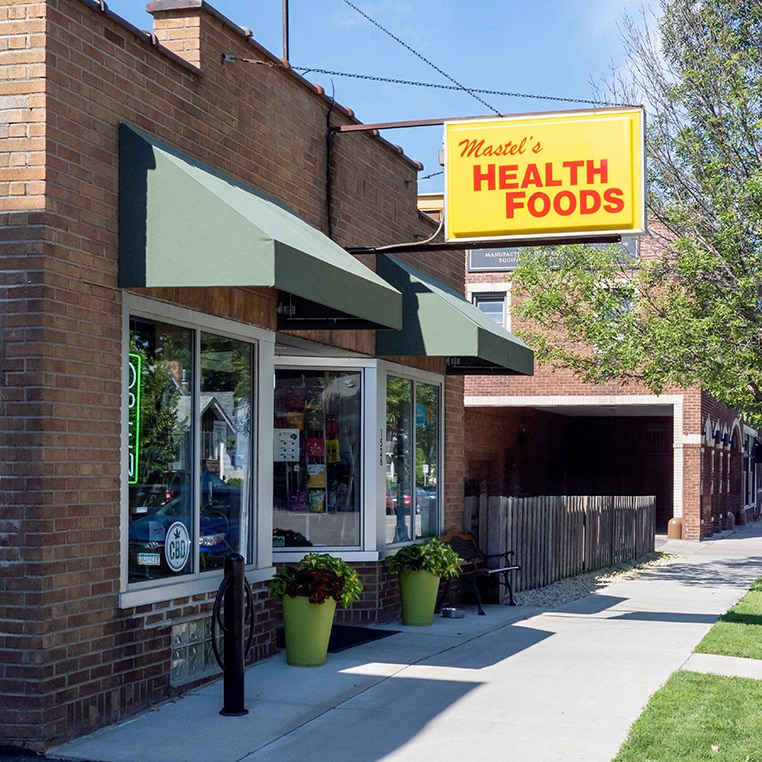 Mastel's Health Foods storefront at 1526 Saint Clair Avenue in Saint Paul — yellow signage, green awnings, and terra-cotta planters with leafy greens by the entrance.