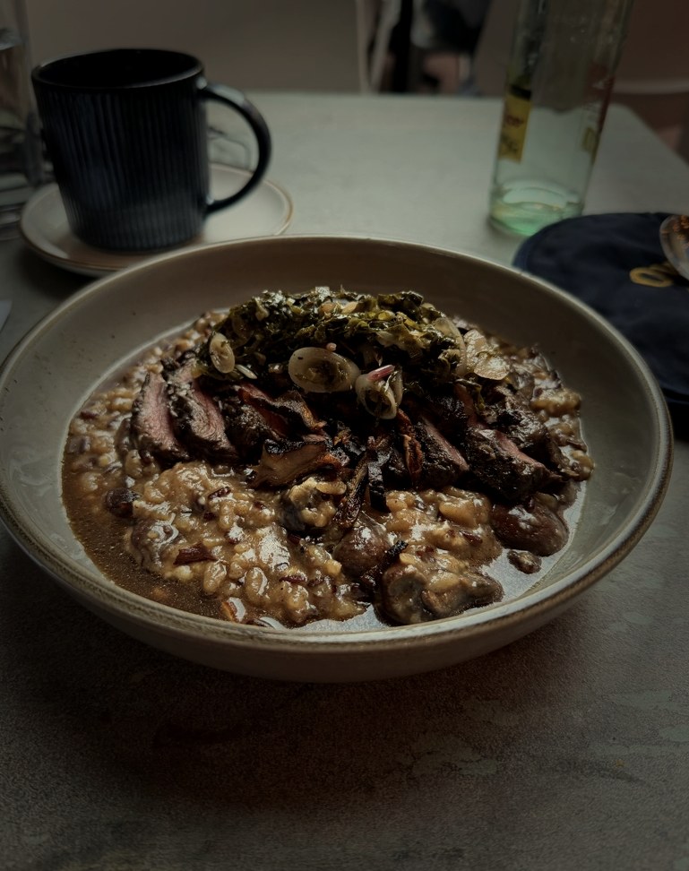 Seared meat with braised greens, mushrooms, and barley risotto in a ceramic bowl at Oro by Nixta — photographed in candlelight.